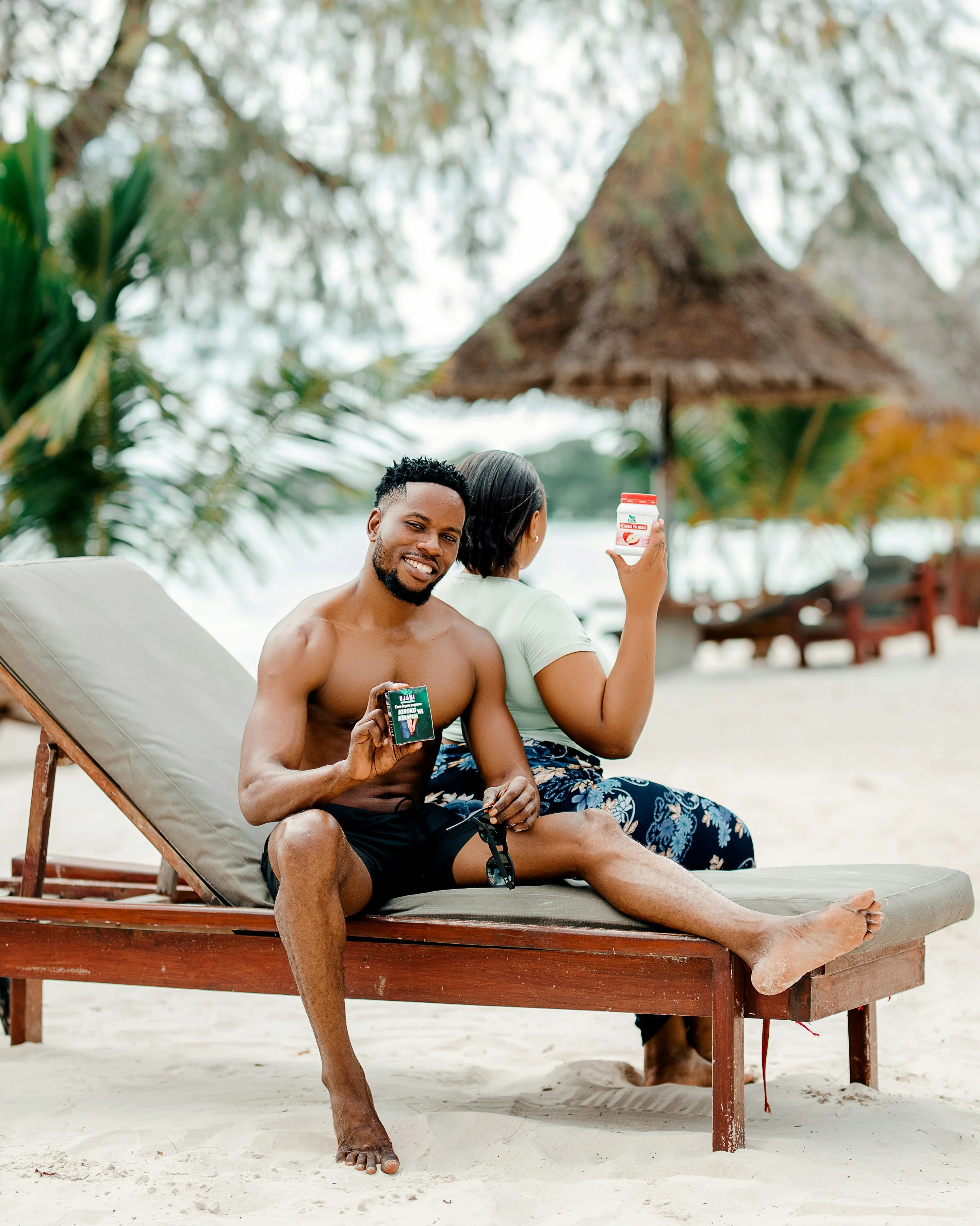 A smiling couple on a wooden lounger on white sand, drinks in hand