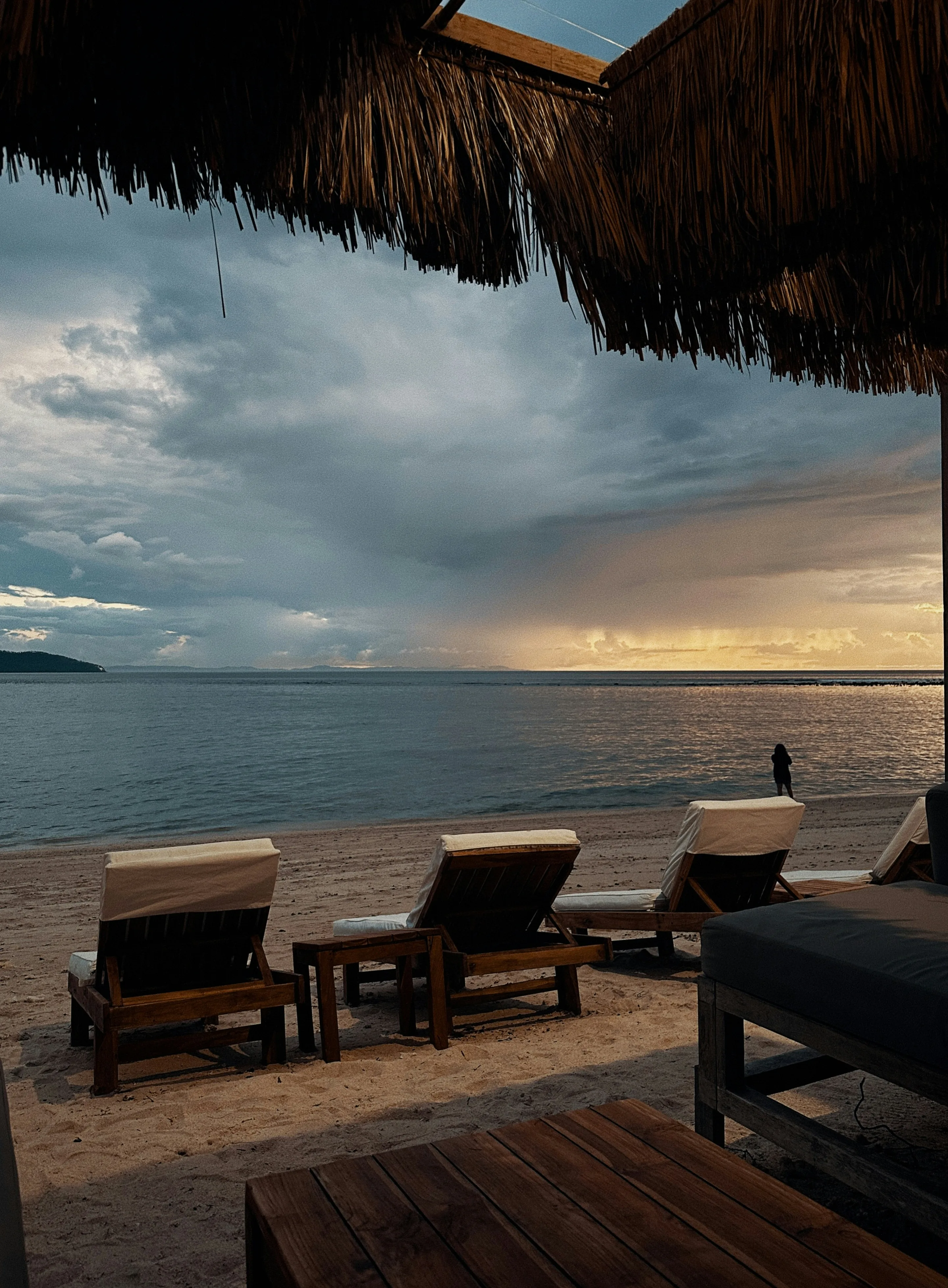 Timber loungers on the sand under a thatched roof, calm water beyond